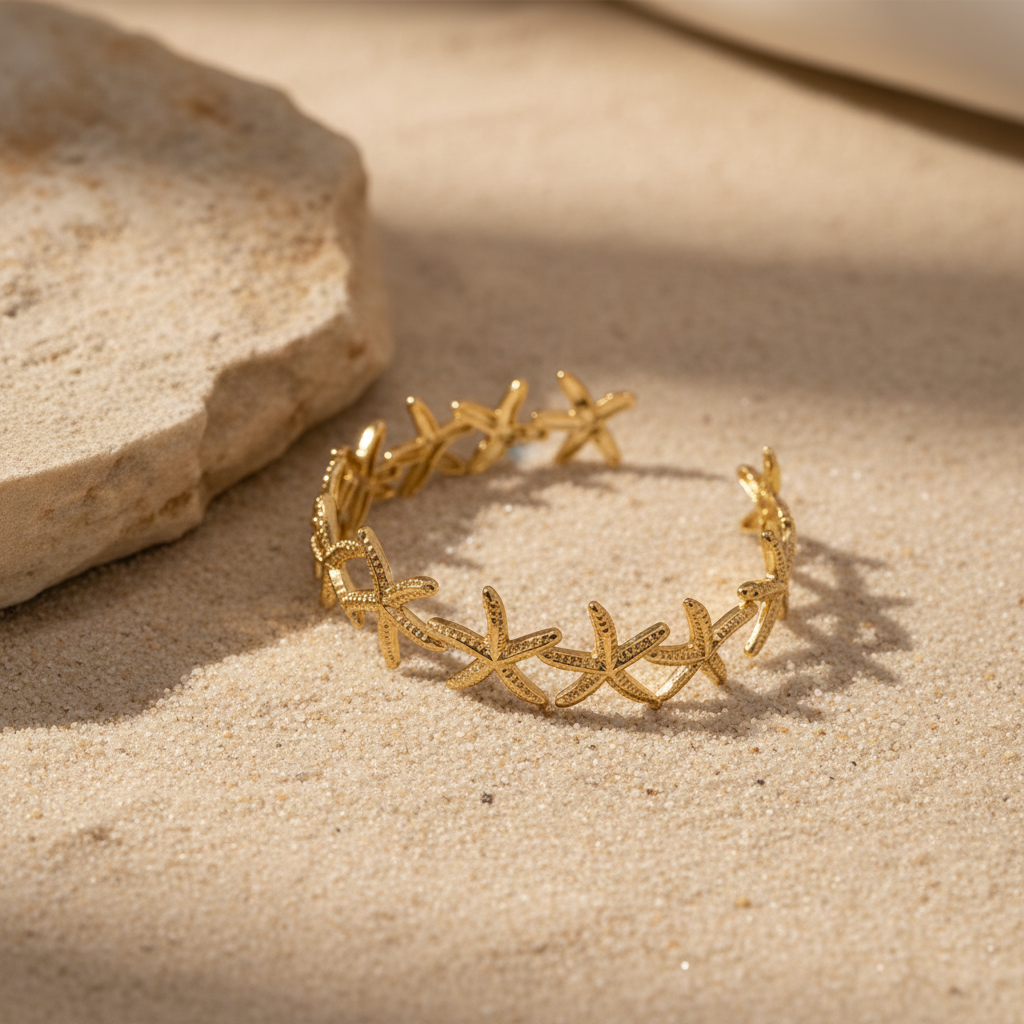 18K gold-plated starfish open cuff bangle angled three-quarter view on beige sandy surface showing full linked starfish design